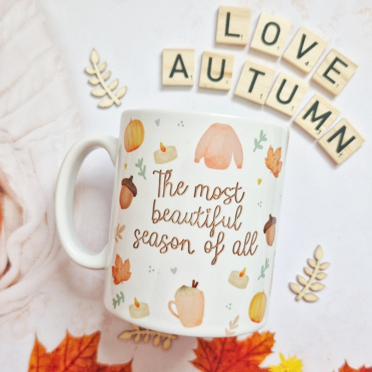A white ceramic mug with the quote "The most beautiful season of all", featuring Autumnal illustrations such as pumpkins, cosy jumpers, acorns, candles and coffee cups