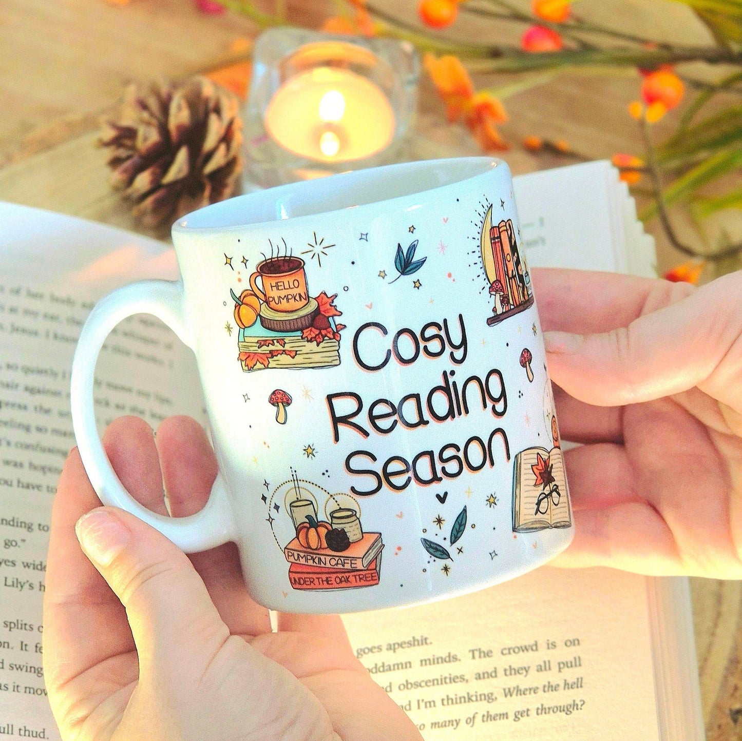 Image shows the same white ceramic mug, with the quote "cosy reading season", surrounded by Autumnal illustrations, being held over an open book in a cosy reading setup.