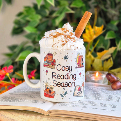 Image shows a white ceramic mug with the quote "Cosy reading season" and autumnal illustrations surrounding it that features books, pumpkins, candles and coffee mugs upon piles of books. The coffee mug is placed upon a book prop, surrounded by autumn leaves and a candle burning and is topped with whipped cream, a chocolate powder dusting and cinnamon stick