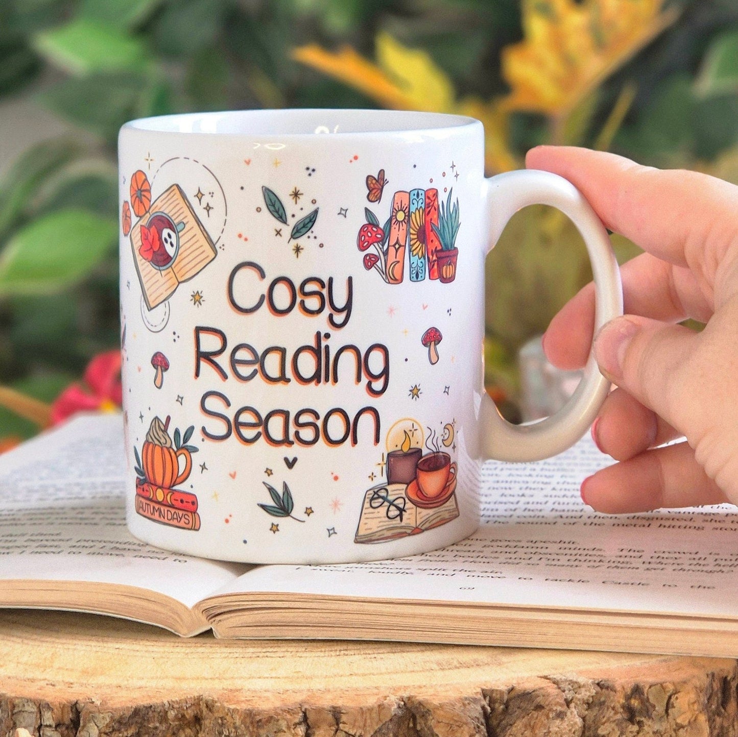 A white ceramic mug with the quote "cosy reading season", placed upon a book with a hand holding it.